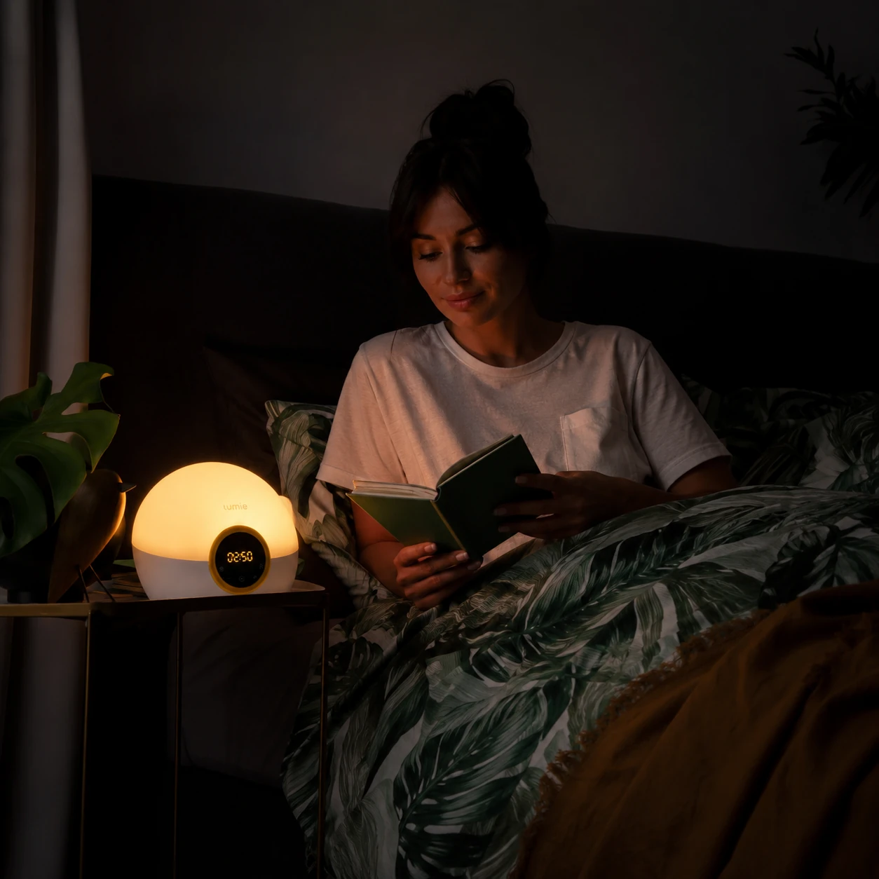 Lumie Bodyclock Rise100 sunrise alarm clock on a bedside table while a woman reads in bed under warm evening light. Lumie Bodyclock Rise100 sunrise alarm clock on a bedside table while a woman reads in bed under warm evening light.